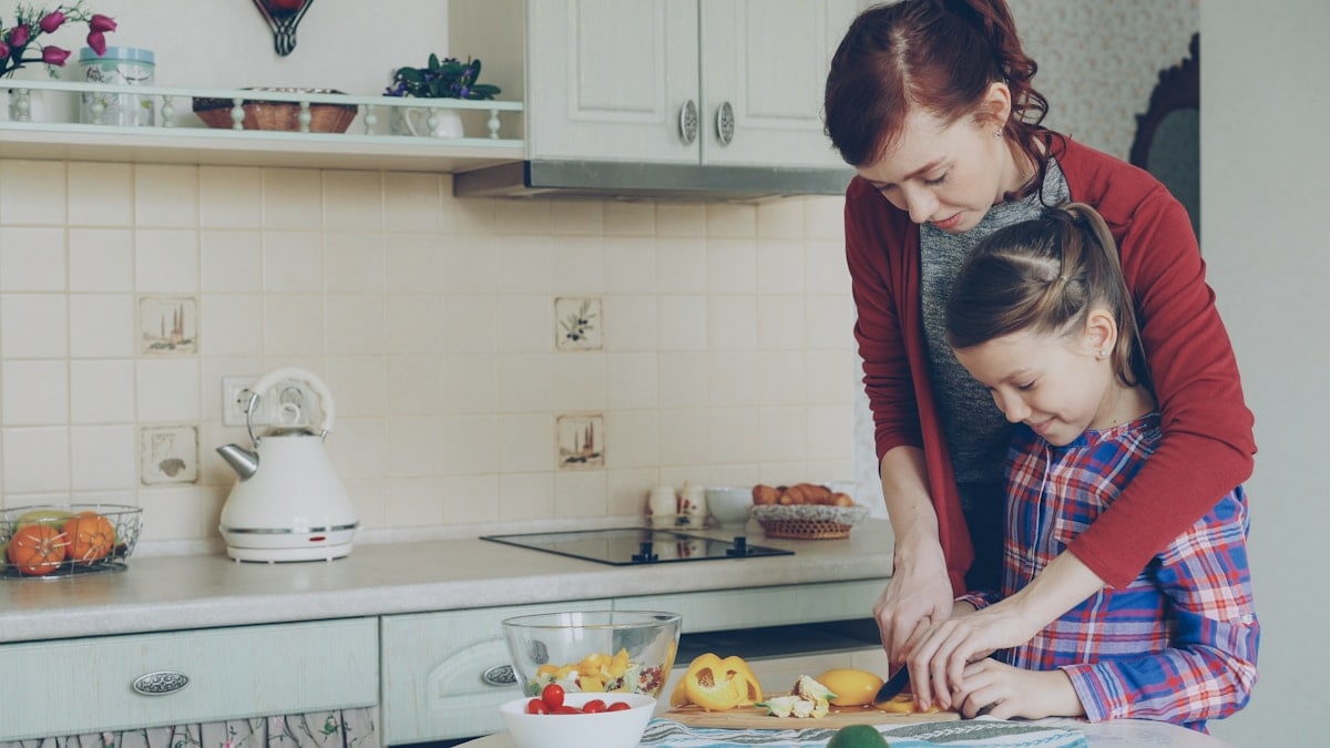 Young pretty mother teaching her cute daughter to cut vegetables properly. Little smiling girl cooking together with loving mom at home in modern kithcen
